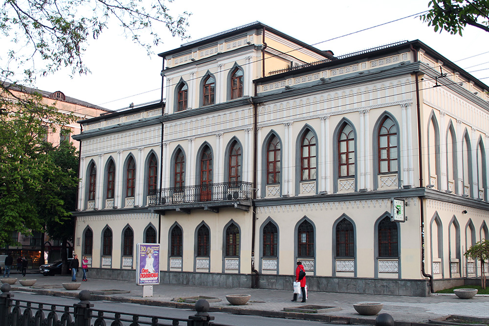Двоповерховий світлий особняк зі стрілчастими вікнами, балконом і підвищеною центральною частиною фасаду