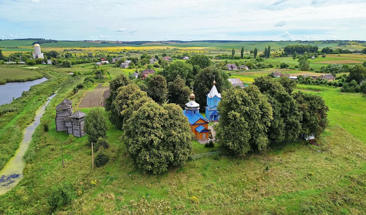 Панорама Пересопниці: дерев’яна церква й окрема дзвіниця серед дерев, ліворуч дві дерев’яні башти сучасної реконструкції, довкола сільські садиби та поля.