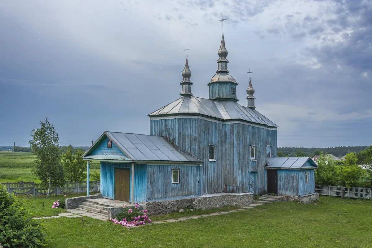 Дерев'яна триверха церква, обшита блакитними дошками, на зеленому подвір'ї під хмарним небом