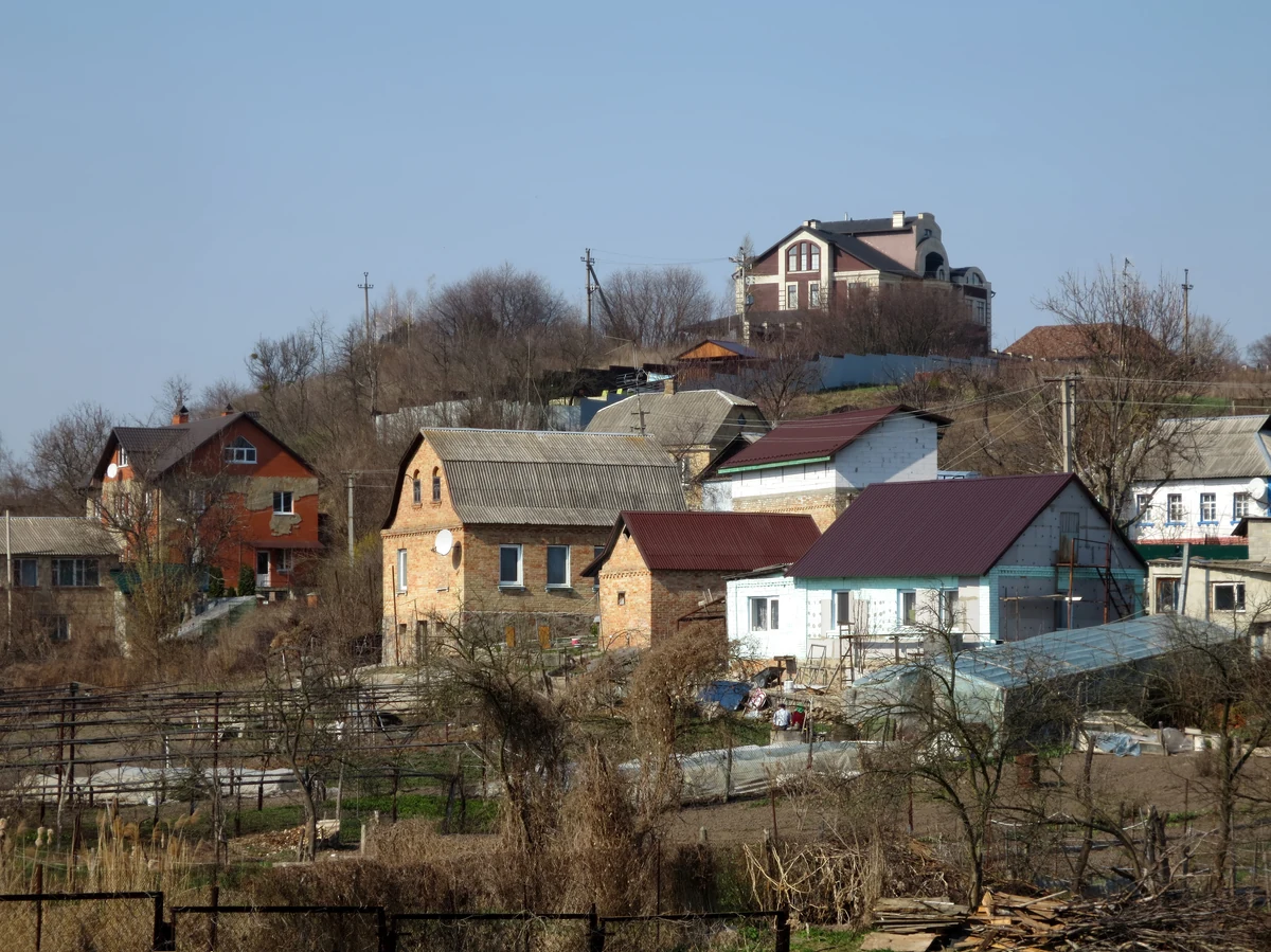 Пагорб із сучасними будинками, огорожами та деревами без листя; на вершині великий будинок із темним дахом, нижче садиби й городи.