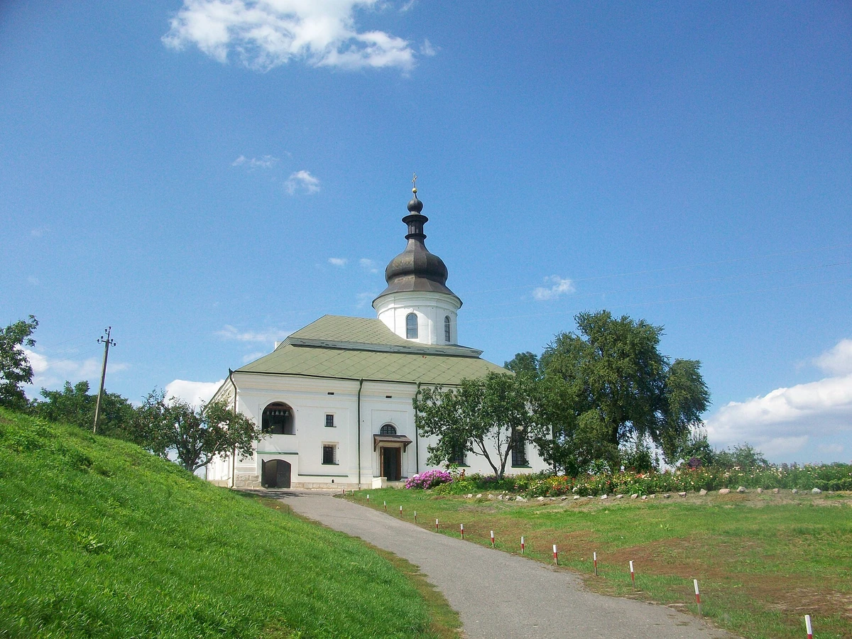 Біла мурована церква із зеленим дахом, темним куполом і дзвонами в арковому отворі; перед будівлею доріжка, трав’яний схил і квітник.