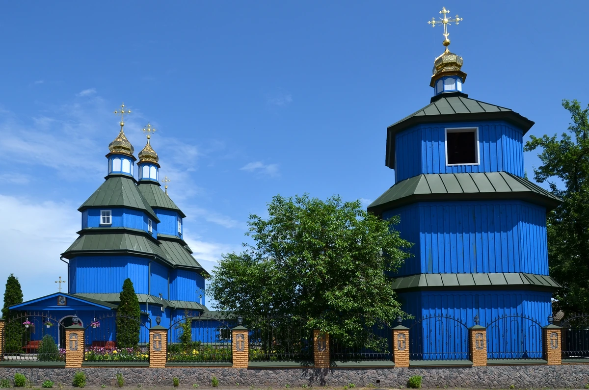 Синяя деревянная церковь слева и колокольня справа за оградой, между ними зелёное дерево.