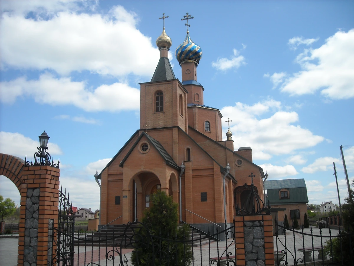 Цегляна церква з банями й хрестами за металевою огорожею під блакитним небом із хмарами.